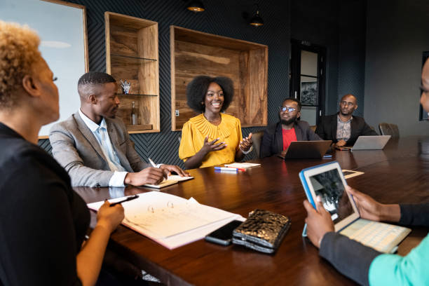 View between colleagues with focus on African professional in early 30s looking across conference table at young associate, smiling and gesturing as she speaks.